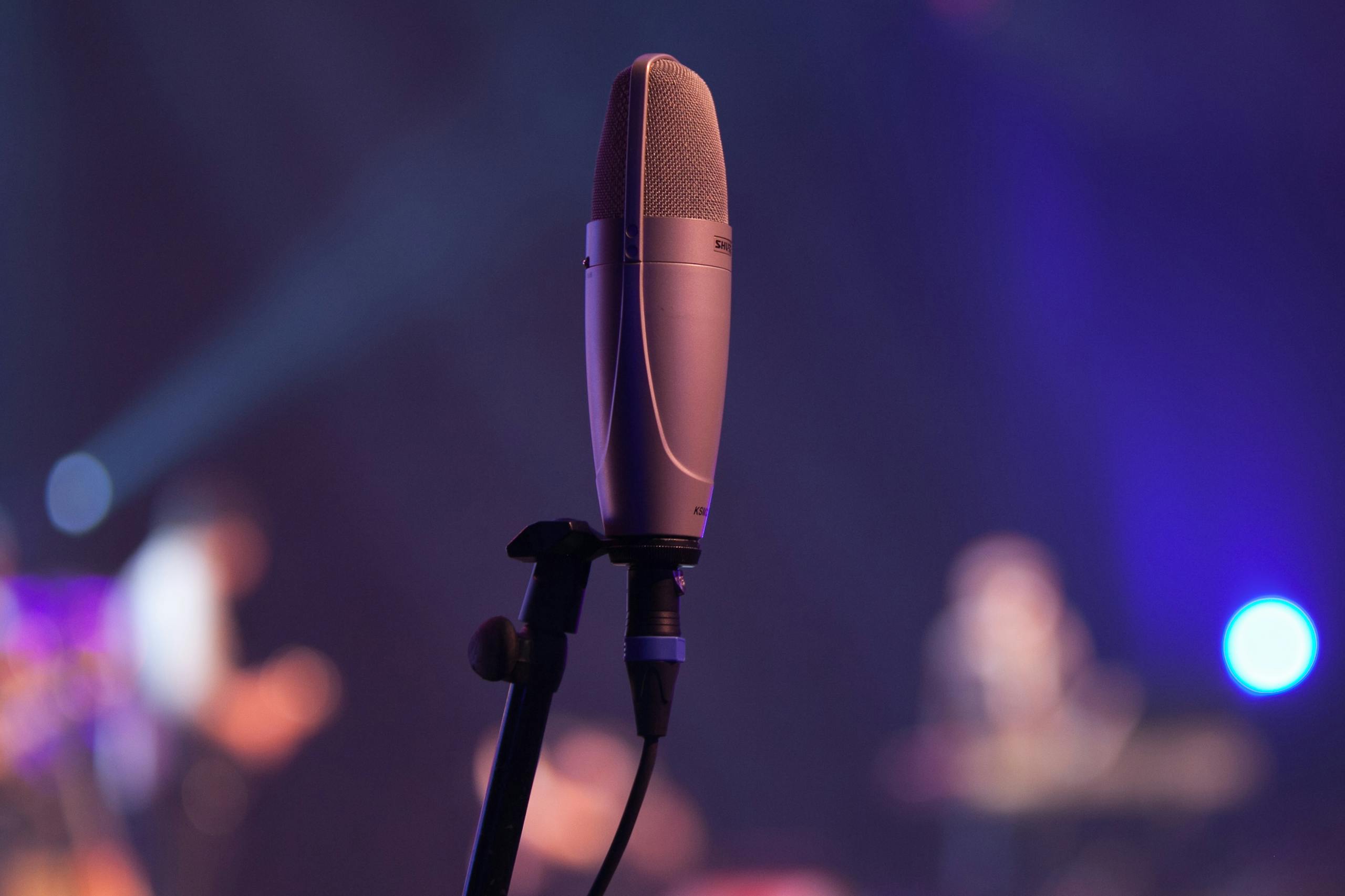 A condenser microphone is highlighted in a music stage setting with a bokeh effect and blurred performers.