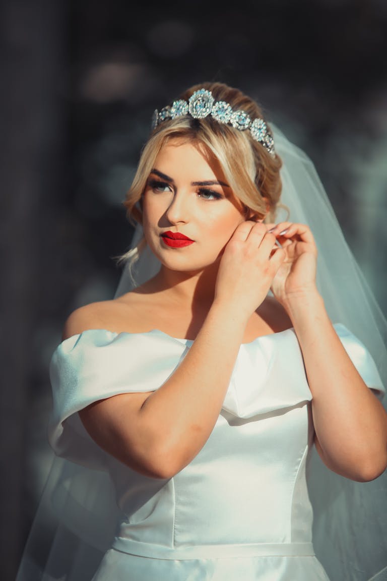 A stylish bride in a white dress and veil adjusts her earring elegantly.