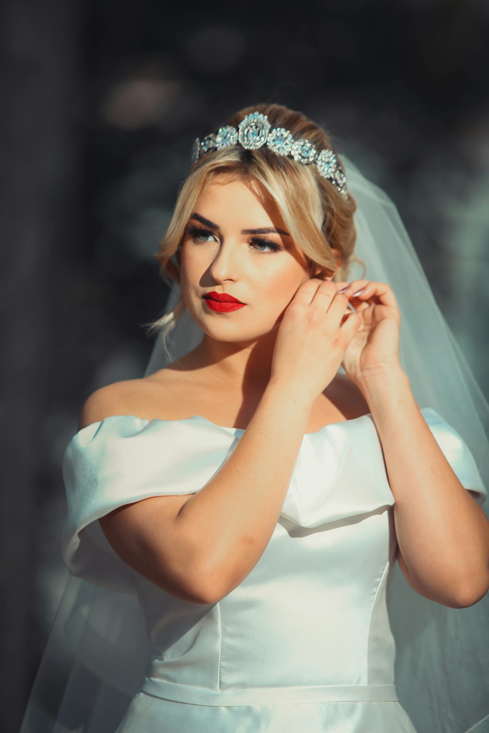 A stylish bride in a white dress and veil adjusts her earring elegantly.