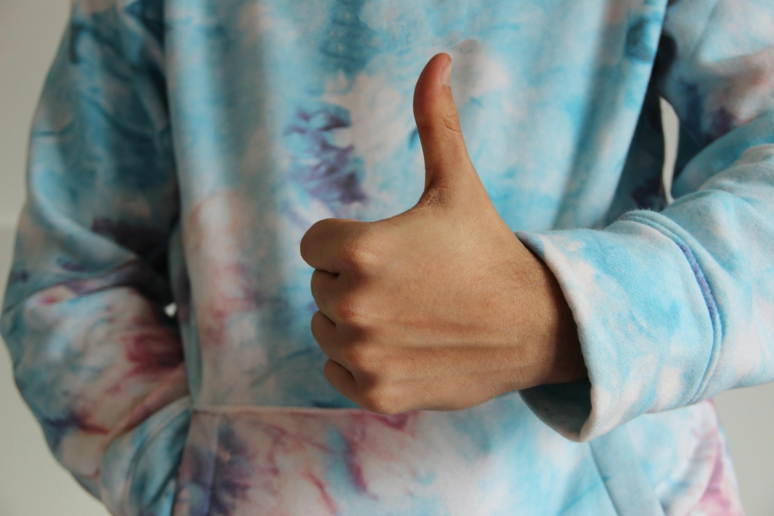 Close-up of a hand giving a thumbs up sign while wearing a colorful tie-dye hoodie.