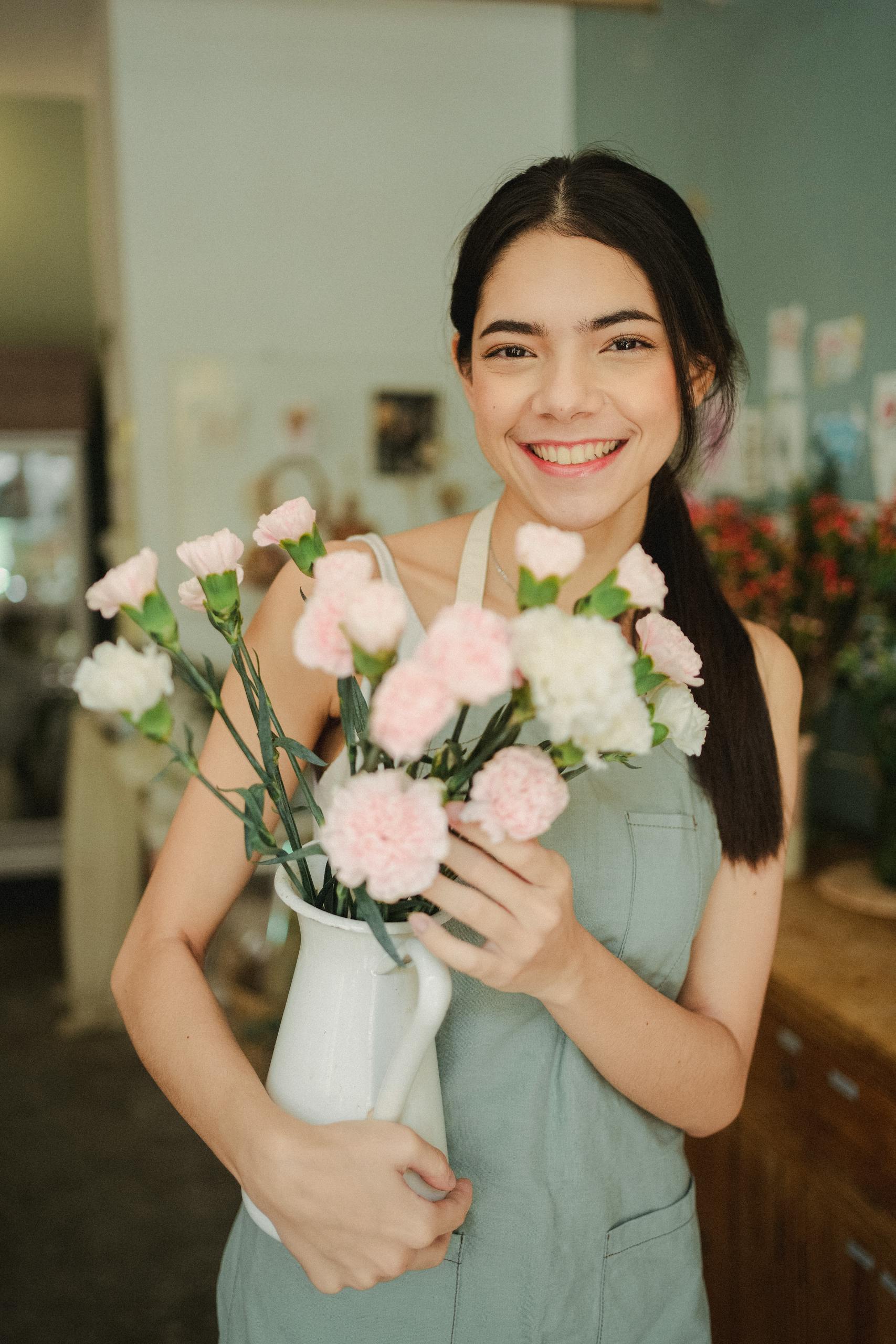 Young female florist in apron holding flowers in vase while smiling and looking at camera